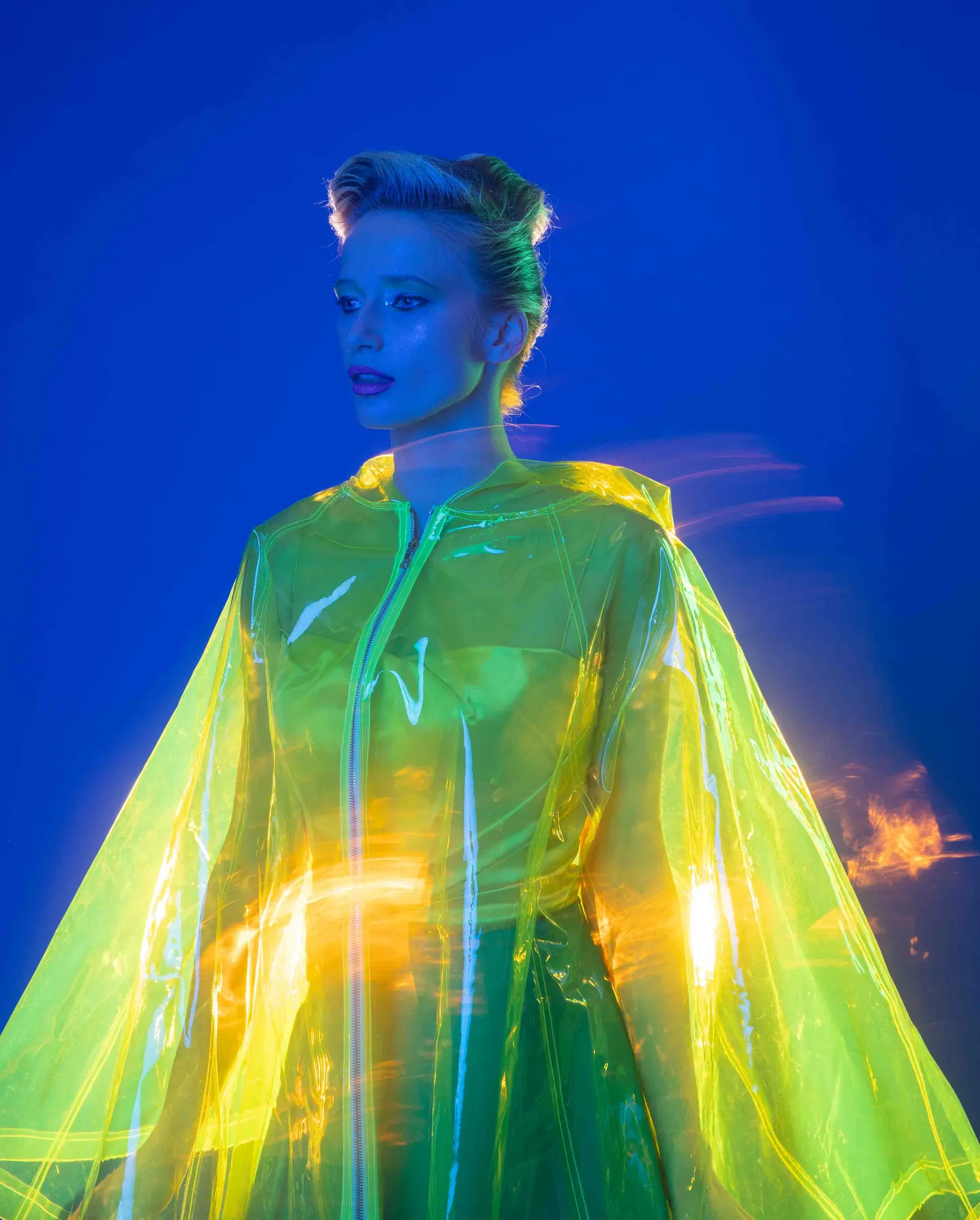 Model in a translucent neon yellow-green cape, lit against a vivid blue backdrop with streaks of motion light