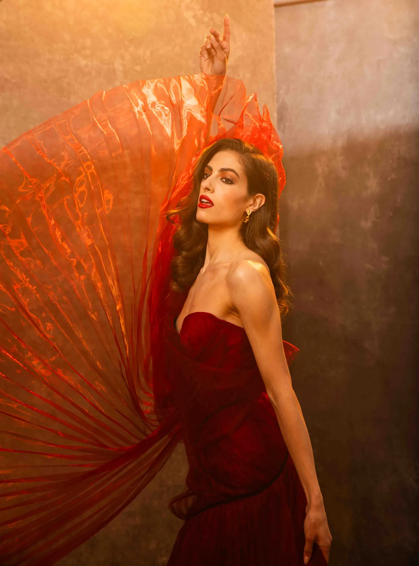 Model in a deep red strapless gown holding a sheer orange pleated fabric like a fan, in warm amber light